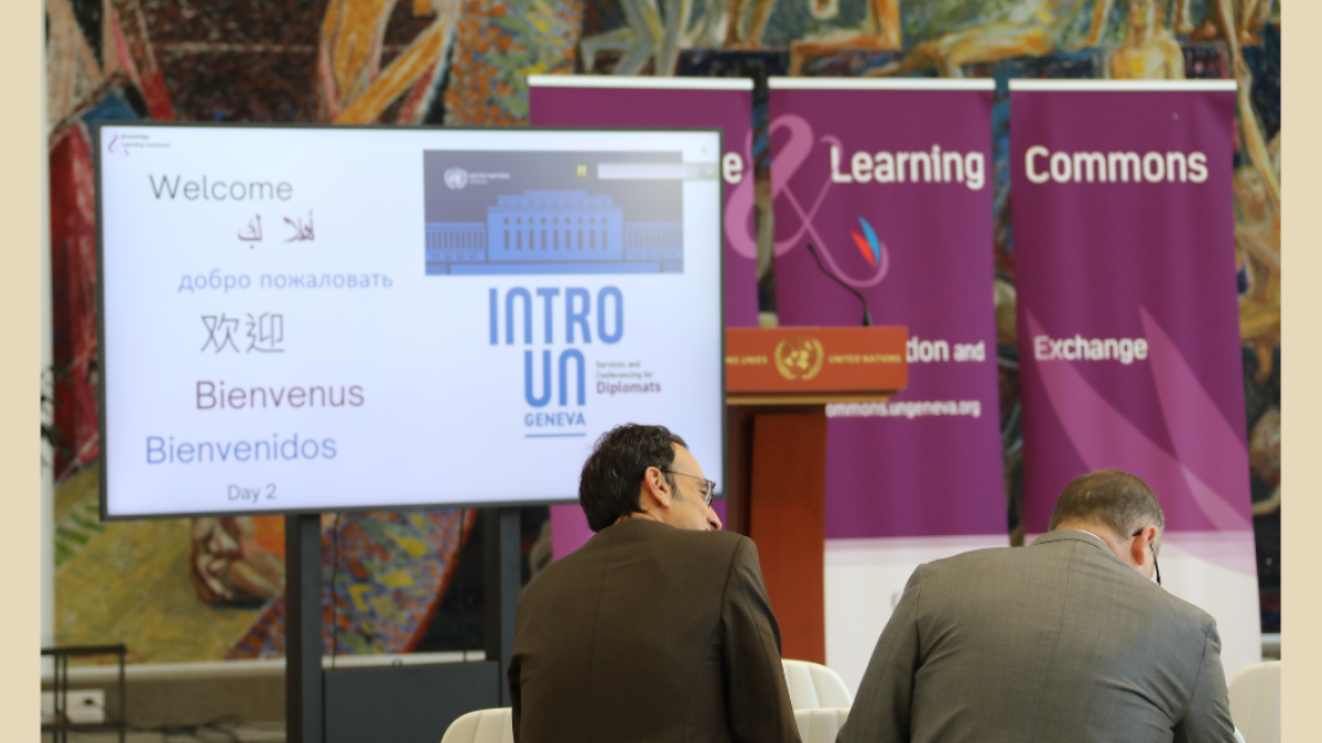 This image shows two diplomats at an event at the UN Library & Archives Geneva, seated and looking at a TV screen featuring the word "Welcome" in several languages.