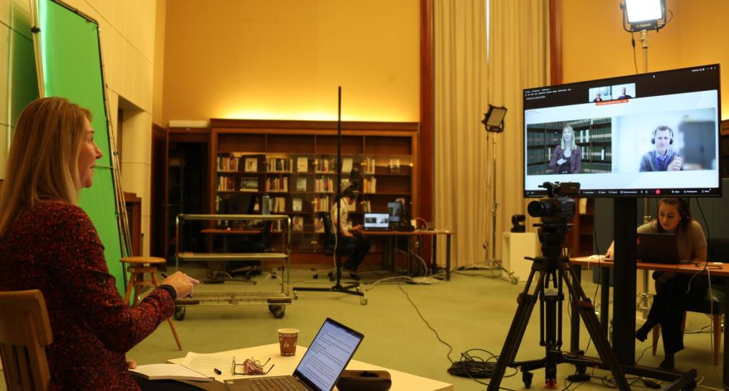 This is a photo from the event on information literacy. It shows the moderator, Sigrun Habermann, looking at a large TV screen, where the other speakers are presenting online. Behind the screen, one of the event producers is working on a laptop. 