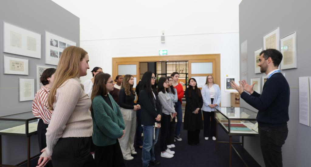 The students visit the UN Museum. This image shows the students looking at materials, on display in the Museum. They are guided by an archivist from the UN Library & Archives Geneva.