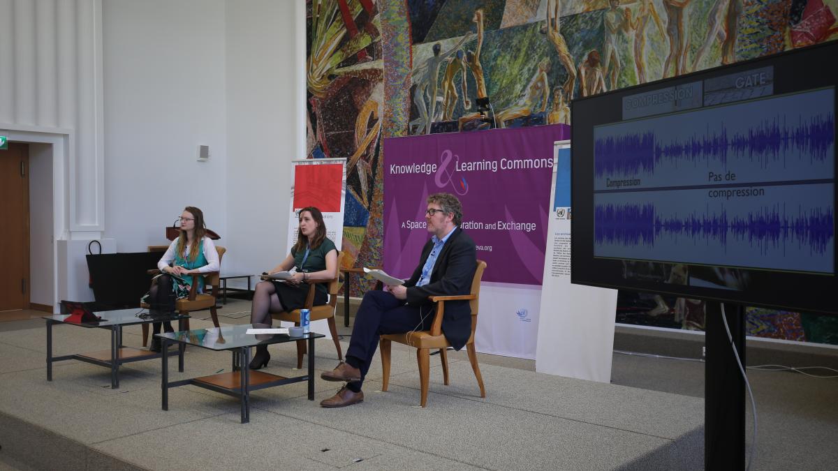 Picture of three people sitting down on a small elevated stage having a interactive conversation. There are two woman and one man. In the background there is a purple board with the Knowledge& Learning Commons logo and as well we can see a tv screen