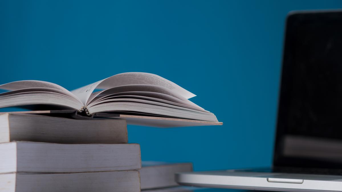 Picture of a couple books stacked with an open book on top and a laptop in the background