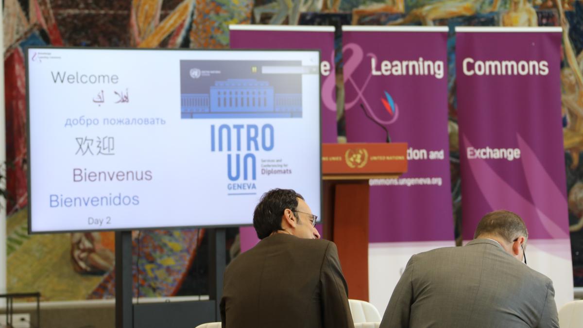 This image shows two diplomats at an event at the UN Library & Archives Geneva, seated and looking at a TV screen featuring the word "Welcome" in several languages.