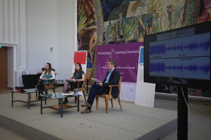 Picture of three people sitting down on a small elevated stage having a interactive conversation. There are two woman and one man. In the background there is a purple board with the Knowledge& Learning Commons logo and as well we can see a tv screen