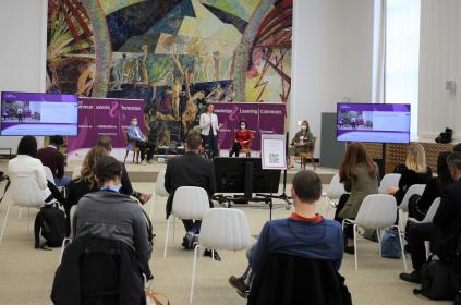 Picture of people sitting in white chairs listening to a woman speak on a small platform satge with two other women sitting next to her