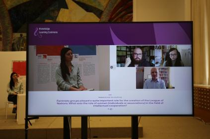 Picture of a TV with four people of the screen having a conversation. In the background we can see the moderator sitting on a chair being filmed for this event. She also appears on the screen