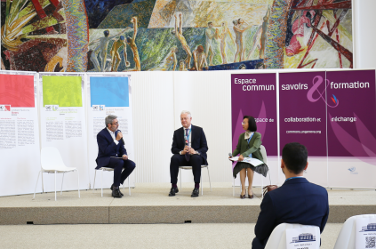 Picture of two men and a woman sitting on the stage platform having a discussion. The background consists of a big white board with posters. 