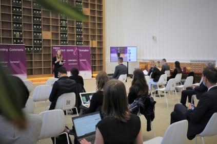 Picture of people sitting in white chairs listening to a woman speak at the front of the room. We can also so a tv with the woman speaking on its screen