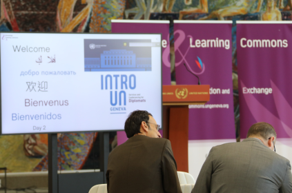 This image shows two diplomats at an event at the UN Library & Archives Geneva, seated and looking at a TV screen featuring the word "Welcome" in several languages.